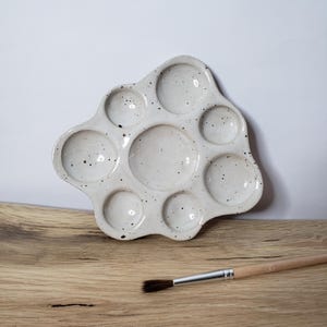 Könnte beinhalten: Eine weiße Keramik-Palette mit acht runden Vertiefungen, die mit schwarzen Punkten übersät sind. Ein Pinsel mit Holzgriff liegt auf einer Holzoberfläche.
