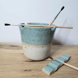 Könnte beinhalten: Ein hellblauer und weißer Keramik-Pinselhalter mit zwei Pinseln darin. Der Halter hat eine gesprenkelte Glasur und eine abgerundete Form. Eine passende hellblaue Keramik-Pinselstütze ist ebenfalls abgebildet.