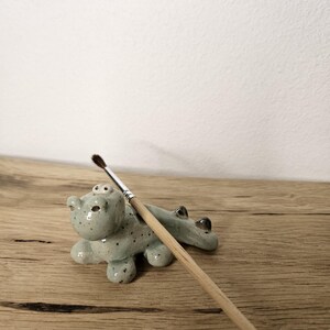Puede incluir: Un soporte para pinceles de cerámica azul claro con forma de oso y motas negras. Un pincel de madera descansa en el soporte. El soporte está sobre una superficie de madera.