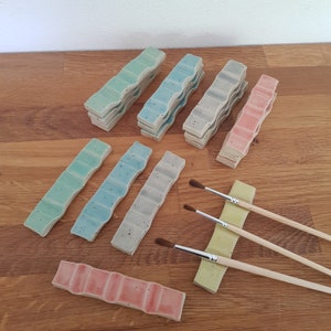 Puede incluir: Un conjunto de 10 paletas de pintura de cerámica en varios tonos de azul, verde y rosa. Las paletas están apiladas y dispuestas sobre una superficie de madera. También se incluyen tres pinceles.