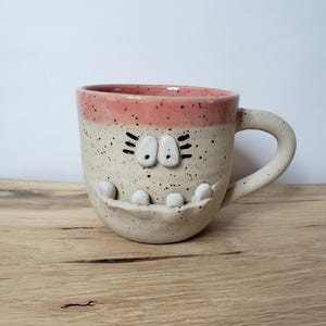 Könnte beinhalten: Eine weiße Keramiktasse mit rosafarbenem Rand und gesprenkeltem Glase. Die Tasse hat ein Gesicht mit weißen Augen, schwarzen Wimpern und einem weißen, zahnigen Grinsen.