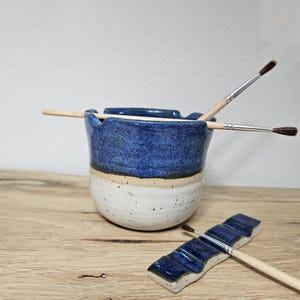 Peut inclure: Un porte-pinceaux en céramique fait à la main avec une base blanche mouchetée, une bande beige et un dessus bleu ondulé. Deux pinceaux sont à l'intérieur et un repose sur un repose-pinceau bleu assorti. Le porte-fournitures d'art est sur une surface en bois.