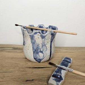 Könnte beinhalten: Ein Keramik-Pinselhalter und Pinselablage-Set. Der Pinselhalter ist weiß mit blauen Tropfen und einem wellenförmigen Rand. Ein Pinsel ruht in der Halterung. Die Pinselablage ist ein kleines, rechteckiges Stück mit blauen Akzenten.