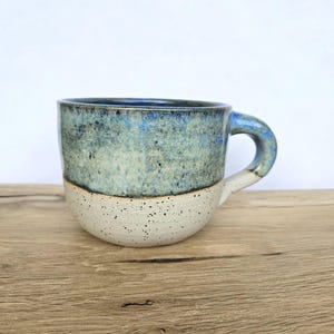 Könnte beinhalten: Ein Keramikbecher mit zweifarbigem Design, bestehend aus einer cremefarbenen Basis mit Sprenkeln und einem strukturierten oberen Bereich in Blau- und Grautönen. Der Becher hat einen runden Körper und einen geschwungenen Henkel mit Glasur.