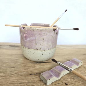 Peut inclure: Un pot à pinceaux en céramique avec un émail moucheté lavande et crème, doté de bords crantés pour maintenir les pinceaux. À côté se trouve un repose-pinceau rectangulaire assorti avec plusieurs rainures, tenant également un pinceau.