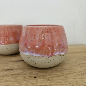Könnte beinhalten: Zwei abgerundete Keramikbecher mit einer glänzenden korallenrosa Glasur im oberen Bereich und einer gesprenkelten, matten cremeweißen Oberfläche an der Basis. Die Becher haben eine bauchige Form mit einem glatten Rand und einem deutlichen Farbübergang.