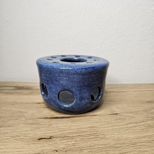May include: A blue ceramic tea warmer with a speckled glaze. The tea warmer has a cylindrical shape with a series of circular holes around the sides and top. The tea warmer is sitting on a wooden surface.