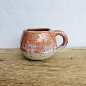May include: A round ceramic mug featuring a glossy coral-pink upper section that transitions into a speckled cream-colored base. The mug has a curved handle and a smooth, glazed finish, showcasing a distinct two-tone color palette and texture.