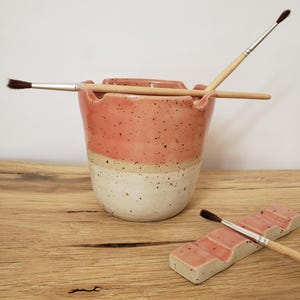 Könnte beinhalten: Ein Set aus zwei Keramik-Pinselhaltern. Der größere Halter ist ein gesprenkelter rosa-weißer Zylinder mit einer breiten Öffnung oben. Der kleinere Halter ist ein gesprenkeltes rosa-weißes Rechteck mit fünf Rillen zum Halten von Pinseln.