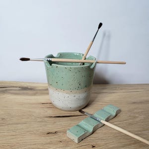 Könnte beinhalten: Zwei Keramik-Pinselhalter. Der größere Halter ist grün und weiß mit Sprenkeln und enthält drei Pinsel. Der kleinere Halter ist grün mit Sprenkeln und enthält einen Pinsel.