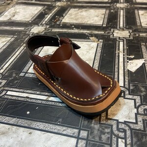 K&ouml;nnte beinhalten: Ein Paar braune Ledersandalen mit einem Riemen und einer Schnalle. Die Sandalen haben einen gen&auml;hten Rand und eine braune Sohle.