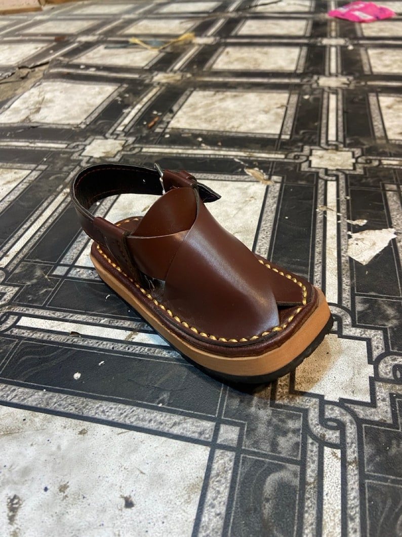 K&ouml;nnte beinhalten: Ein Paar braune Ledersandalen mit einem Riemen und einer Schnalle. Die Sandalen haben einen gen&auml;hten Rand und eine braune Sohle.