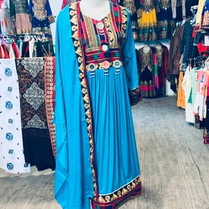 K&ouml;nnte beinhalten: Ein lebendiges t&uuml;rkisfarbenes Kleid mit aufwendigen Stickereien und Verzierungen. Das Kleid hat einen V-Ausschnitt, lange &Auml;rmel und einen flie&szlig;enden Rock. Die Stickerei enth&auml;lt rote, goldene und schwarze Details. Ein passender t&uuml;rkisfarbener Schal ist &uuml;ber dem Kleid drapiert. Weitere Kleider sind im Hintergrund zu sehen.