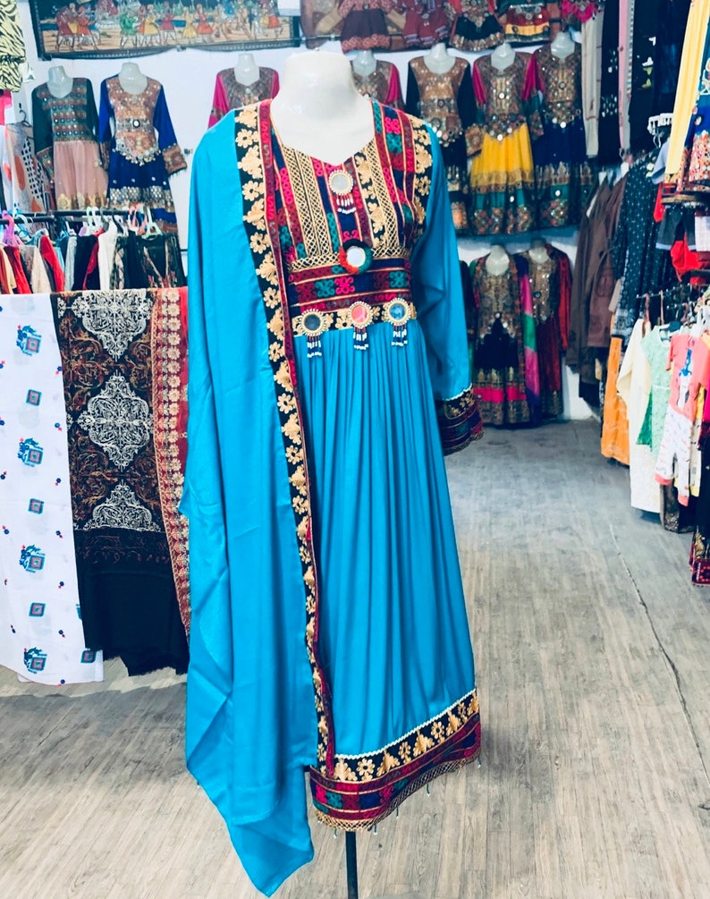 K&ouml;nnte beinhalten: Ein lebendiges t&uuml;rkisfarbenes Kleid mit aufwendigen Stickereien und Verzierungen. Das Kleid hat einen V-Ausschnitt, lange &Auml;rmel und einen flie&szlig;enden Rock. Die Stickerei enth&auml;lt rote, goldene und schwarze Details. Ein passender t&uuml;rkisfarbener Schal ist &uuml;ber dem Kleid drapiert. Weitere Kleider sind im Hintergrund zu sehen.