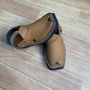 Op de afbeelding: Een paar bruine leren sandalen met een zwarte zool en witte stiksels. De sandalen hebben een enkelband en een gesp. Het leer heeft een textuur. De sandalen zijn van leer.