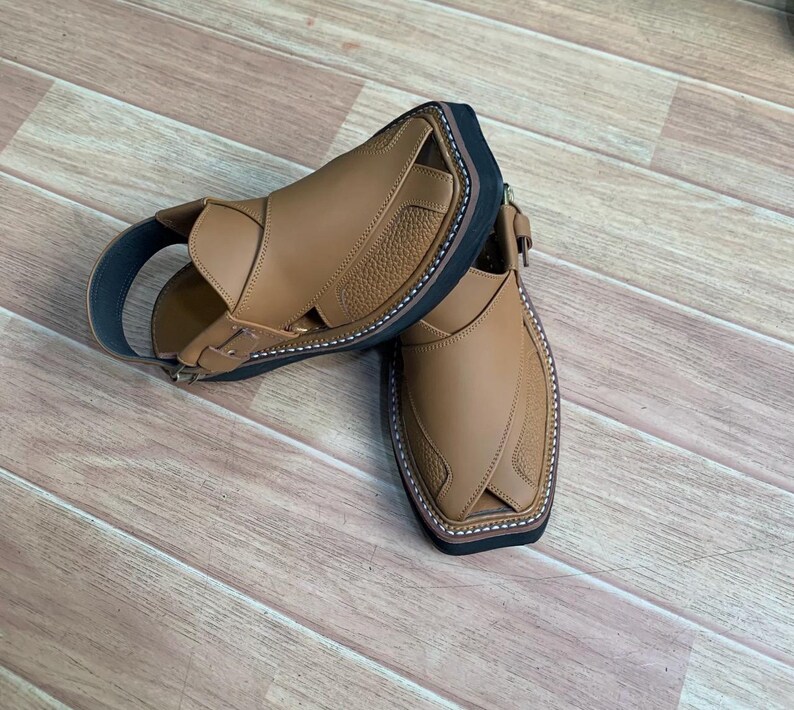 Op de afbeelding: Een paar bruine leren sandalen met een zwarte zool en witte stiksels. De sandalen hebben een enkelband en een gesp. Het leer heeft een textuur. De sandalen zijn van leer.