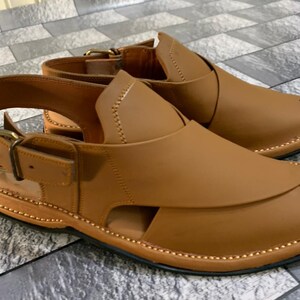 May include: A pair of brown leather sandals with a buckle strap and a decorative stitching detail around the edge.