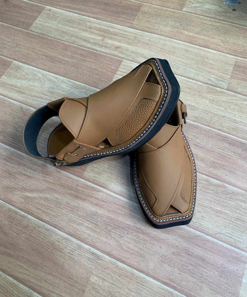 Op de afbeelding: Een paar bruinleren sandalen met een zwarte zool en band. De sandalen hebben een textuurontwerp op de bovenkant en zijn gestikt met witte draad. De sandalen worden gepresenteerd op een houten oppervlak.