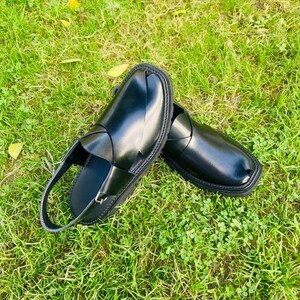 May include: A pair of black leather sandals with a strap across the top and a buckle closure. The sandals are on a green grassy surface.