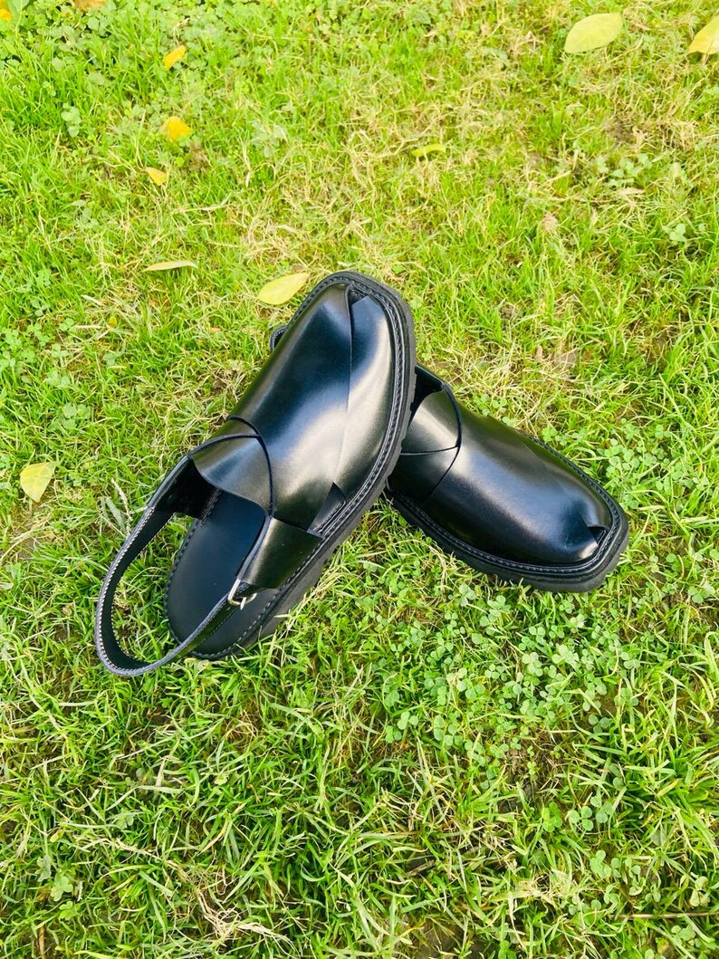 May include: A pair of black leather sandals with a strap across the top and a buckle closure. The sandals are on a green grassy surface.