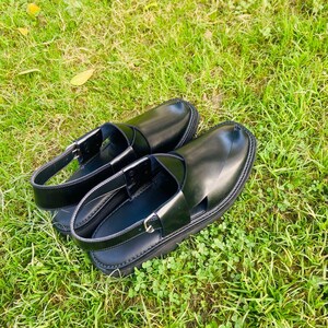 May include: A pair of black leather sandals with a strap across the top and an open toe design. The sandals are sitting on a bed of green grass.