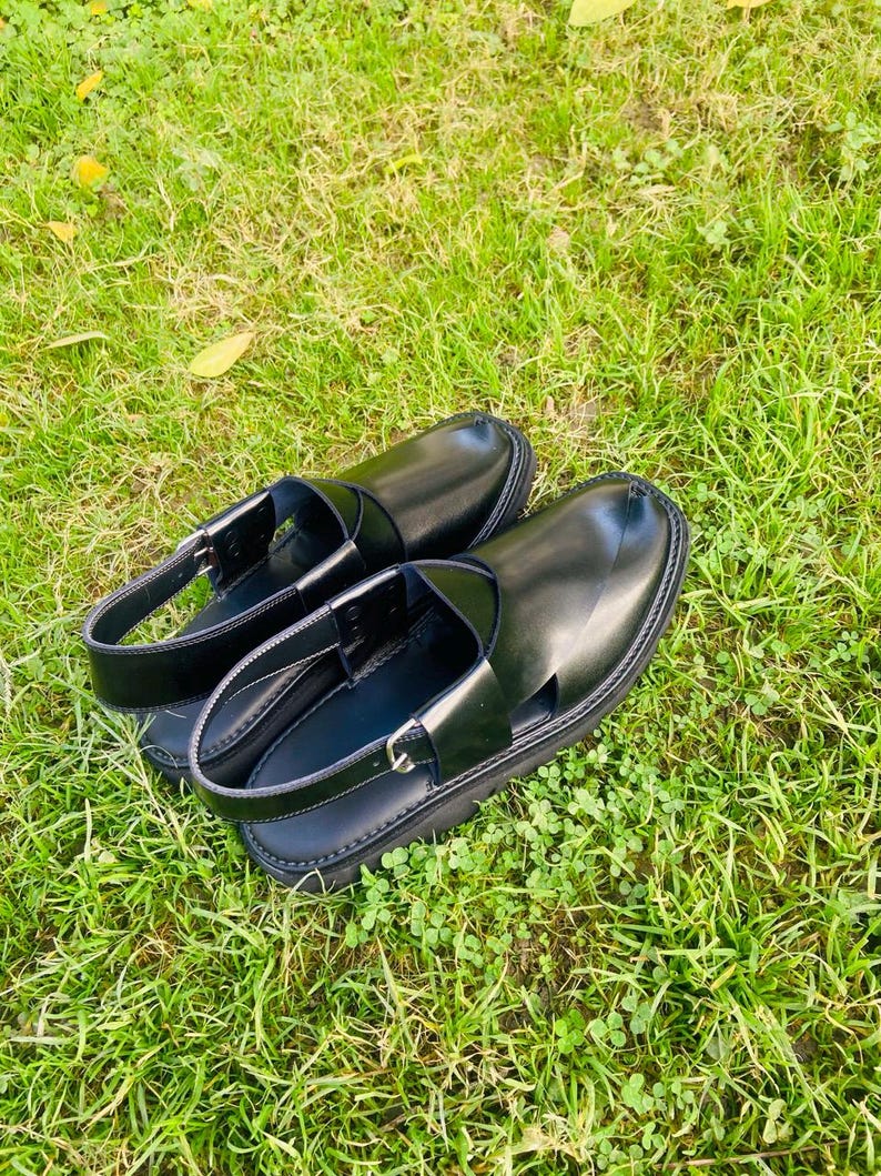 May include: A pair of black leather sandals with a strap across the top and an open toe design. The sandals are sitting on a bed of green grass.