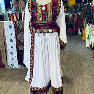 K&ouml;nnte beinhalten: Ein wei&szlig;es Kleid mit langen &Auml;rmeln und einem farbenfrohen bestickten Oberteil und G&uuml;rtel. Die Stickerei weist rote, gr&uuml;ne, blaue und goldene Muster auf. Das Kleid wird auf einer Schaufensterpuppe in einem Gesch&auml;ft pr&auml;sentiert, mit anderen Kleidern im Hintergrund.