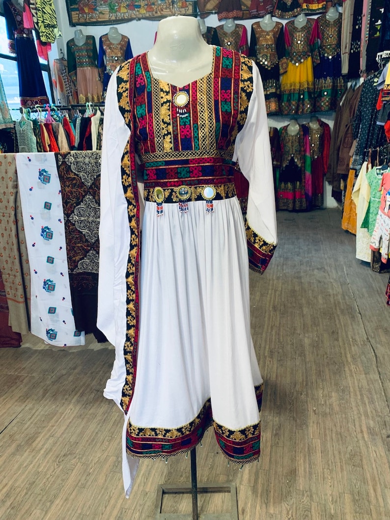 K&ouml;nnte beinhalten: Ein wei&szlig;es Kleid mit langen &Auml;rmeln und einem farbenfrohen bestickten Oberteil und G&uuml;rtel. Die Stickerei weist rote, gr&uuml;ne, blaue und goldene Muster auf. Das Kleid wird auf einer Schaufensterpuppe in einem Gesch&auml;ft pr&auml;sentiert, mit anderen Kleidern im Hintergrund.