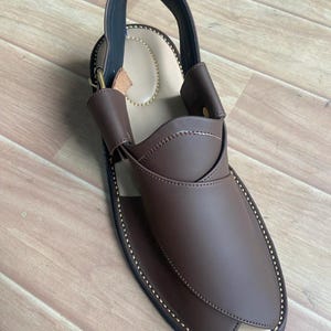 以下が含まれることがあります： ストラップとバックルが付いた茶色の革サンダル。サンダルはつま先が尖っており、縁にステッチが施されています。