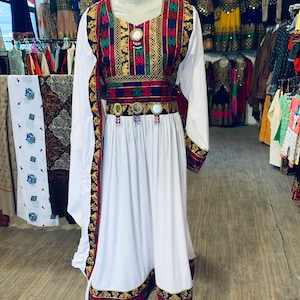 K&ouml;nnte beinhalten: Ein wei&szlig;es Kleid mit langen &Auml;rmeln und farbenfrohen Stickereien. Das Kleid hat einen quadratischen Ausschnitt und einen dekorativen G&uuml;rtel mit Spiegelakzenten. Die Manschetten und der Saum sind mit passenden Stickereien verziert. Das Kleid wird auf einer Schaufensterpuppe pr&auml;sentiert.