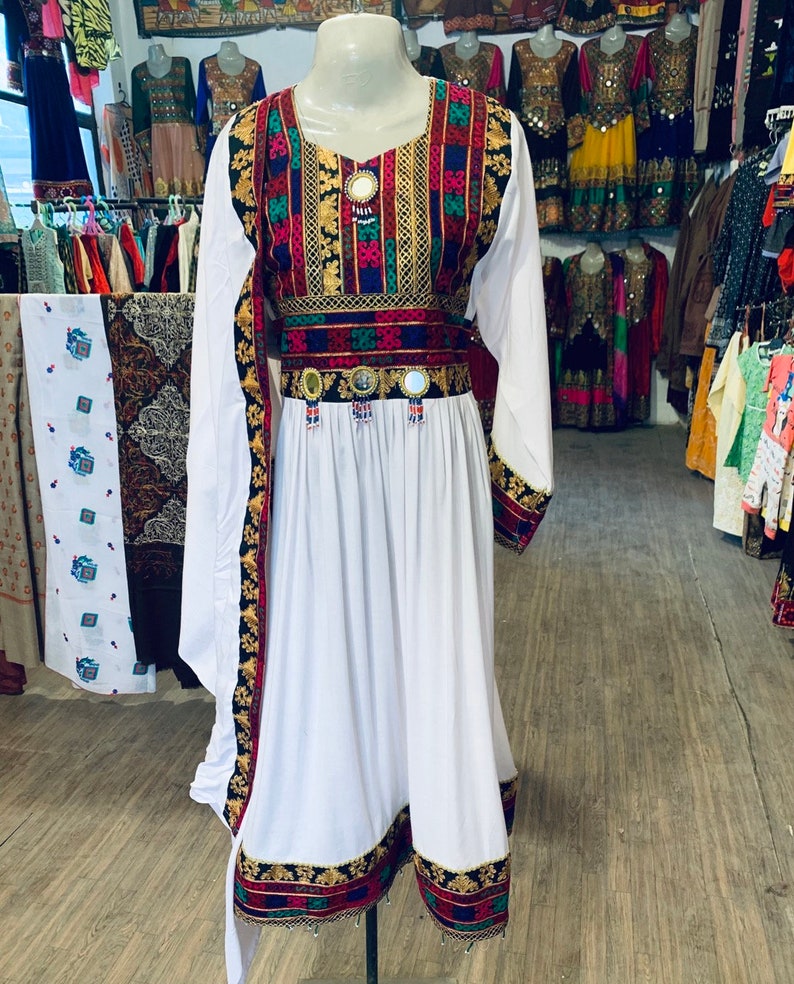 K&ouml;nnte beinhalten: Ein wei&szlig;es Kleid mit langen &Auml;rmeln und farbenfrohen Stickereien. Das Kleid hat einen quadratischen Ausschnitt und einen dekorativen G&uuml;rtel mit Spiegelakzenten. Die Manschetten und der Saum sind mit passenden Stickereien verziert. Das Kleid wird auf einer Schaufensterpuppe pr&auml;sentiert.