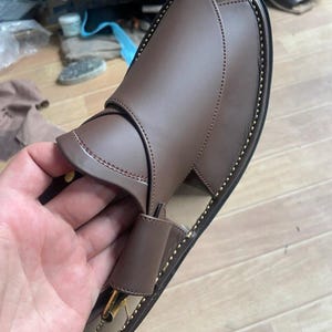 以下が含まれることがあります： 金色の縁取りとステッチが施された茶色の革製のサンダル。サンダルは足首に巻き付けるストラップとつま先ストラップが付いています。