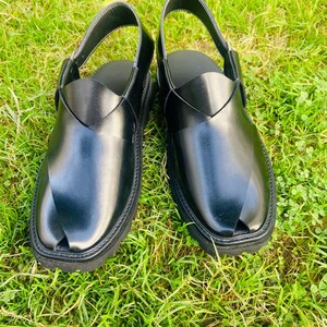 May include: A pair of black leather sandals with a strap across the top and a pointed toe. The sandals are on a green grassy surface.
