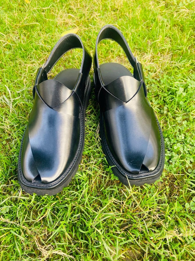 May include: A pair of black leather sandals with a strap across the top and a pointed toe. The sandals are on a green grassy surface.