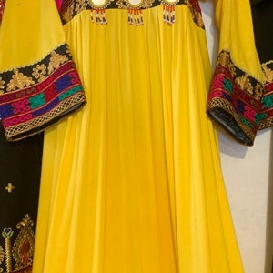 K&ouml;nnte beinhalten: Ein langes, flie&szlig;endes gelbes Kleid mit aufwendiger Stickerei und Spiegelverzierungen. Das Kleid hat einen quadratischen Ausschnitt und lange &Auml;rmel mit detaillierten Manschetten. Das Oberteil und die &Auml;rmel sind mit farbenfrohen Mustern verziert, auf einer Schaufensterpuppe.