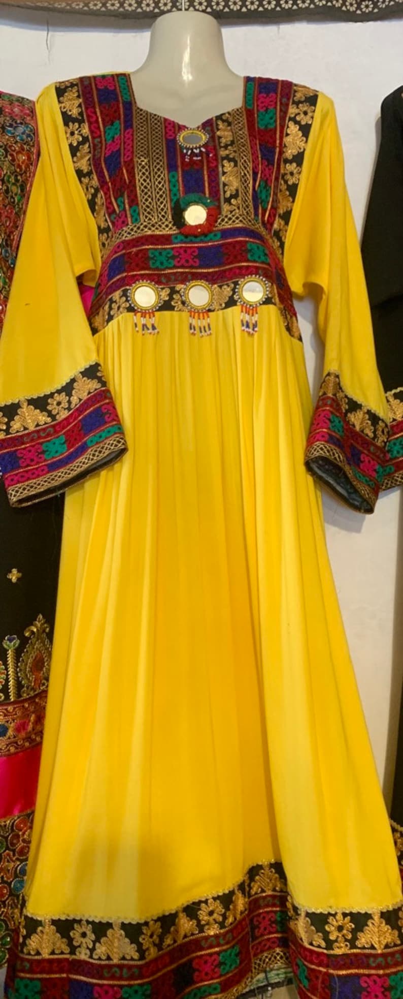 K&ouml;nnte beinhalten: Ein langes, flie&szlig;endes gelbes Kleid mit aufwendiger Stickerei und Spiegelverzierungen. Das Kleid hat einen quadratischen Ausschnitt und lange &Auml;rmel mit detaillierten Manschetten. Das Oberteil und die &Auml;rmel sind mit farbenfrohen Mustern verziert, auf einer Schaufensterpuppe.