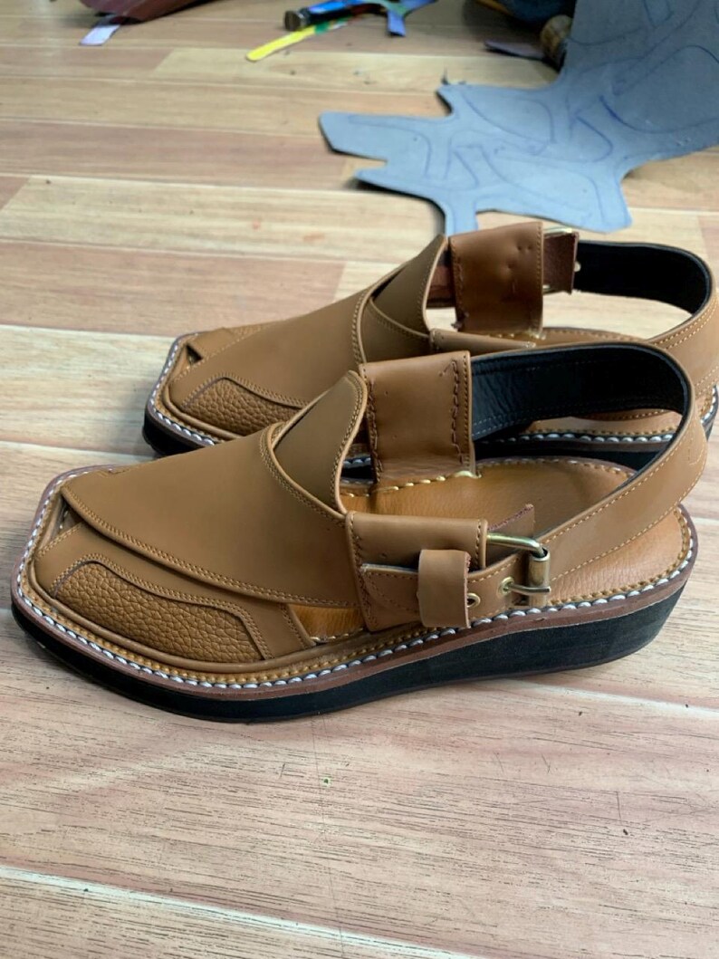 Op de afbeelding: Een paar bruinleren sandalen met een vierkante neus. De sandalen hebben een getextureerde wreef, een gespsluiting en een zwarte zool met witte stiksels. Het schoeisel staat op een houten oppervlak.