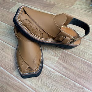 Op de afbeelding: Een paar bruin lederen sandalen met een zwarte zool en een gespsluiting. De sandalen hebben een textuurontwerp op de bovenkant en witte stiksels rond de randen. Gepresenteerd op een houten oppervlak.