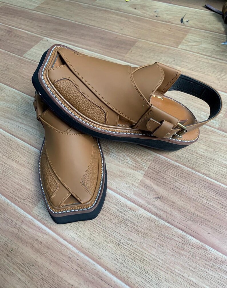 Op de afbeelding: Een paar bruin lederen sandalen met een zwarte zool en een gespsluiting. De sandalen hebben een textuurontwerp op de bovenkant en witte stiksels rond de randen. Gepresenteerd op een houten oppervlak.