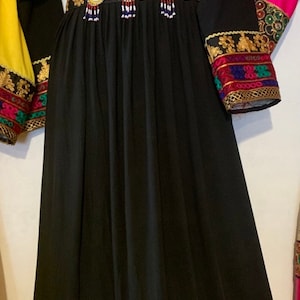 K&ouml;nnte beinhalten: Schwarzes Kleid mit langen &Auml;rmeln und aufwendiger Stickerei. Das Kleid hat goldene, rote, blaue und gr&uuml;ne Akzente am Ausschnitt, an der Taille und an den Manschetten. Der Rock ist aus flie&szlig;endem, einfarbigem schwarzem Stoff. Das Kleid wird auf einer Schaufensterpuppe pr&auml;sentiert.