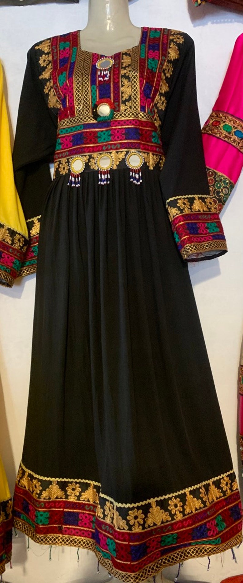 K&ouml;nnte beinhalten: Schwarzes Kleid mit langen &Auml;rmeln und aufwendiger Stickerei. Das Kleid hat goldene, rote, blaue und gr&uuml;ne Akzente am Ausschnitt, an der Taille und an den Manschetten. Der Rock ist aus flie&szlig;endem, einfarbigem schwarzem Stoff. Das Kleid wird auf einer Schaufensterpuppe pr&auml;sentiert.