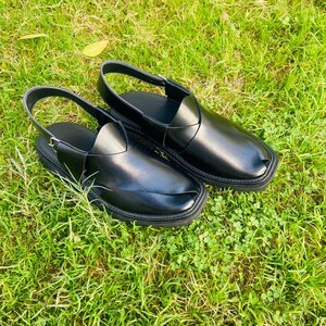 May include: A pair of black leather sandals with a strap across the top and a buckle closure. The sandals are on a green grassy surface.
