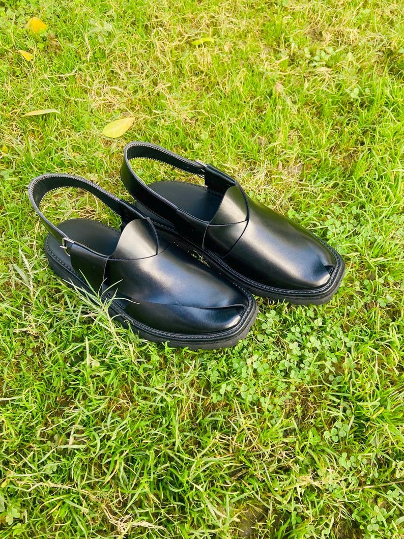 May include: A pair of black leather sandals with a strap across the top and a buckle closure. The sandals are on a green grassy surface.