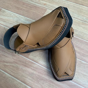 Op de afbeelding: Een paar bruinleren sandalen met een getextureerd bovenwerk en een zwarte zool. De sandalen hebben een enkelband en zijn gestikt met witte draad. Ze worden gepresenteerd op een houten oppervlak.