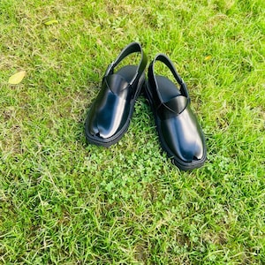 May include: A pair of black leather sandals with a strap across the top of the foot. The sandals are on a green grassy surface.
