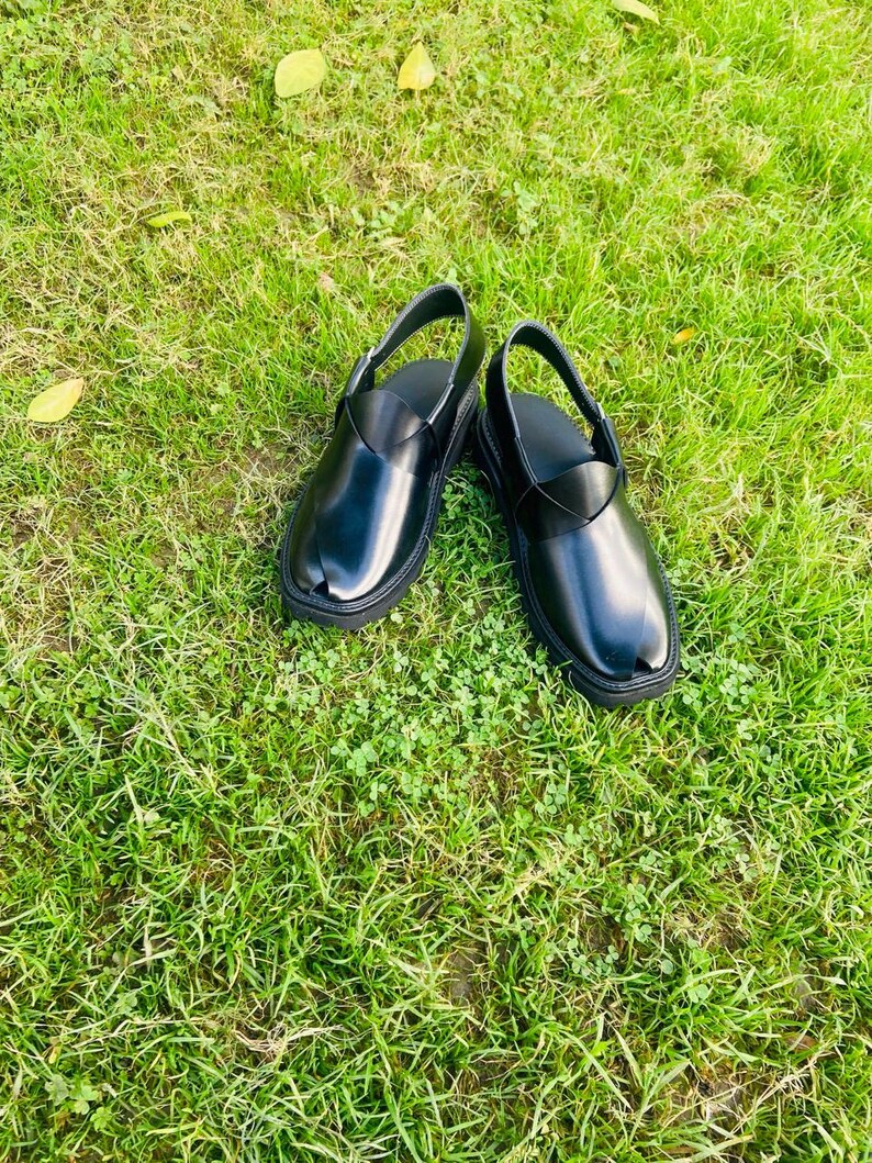 May include: A pair of black leather sandals with a strap across the top of the foot. The sandals are on a green grassy surface.