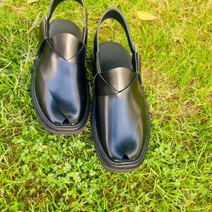 May include: A pair of black leather sandals with a strap across the top and a pointed toe. The sandals are on a green grassy surface.