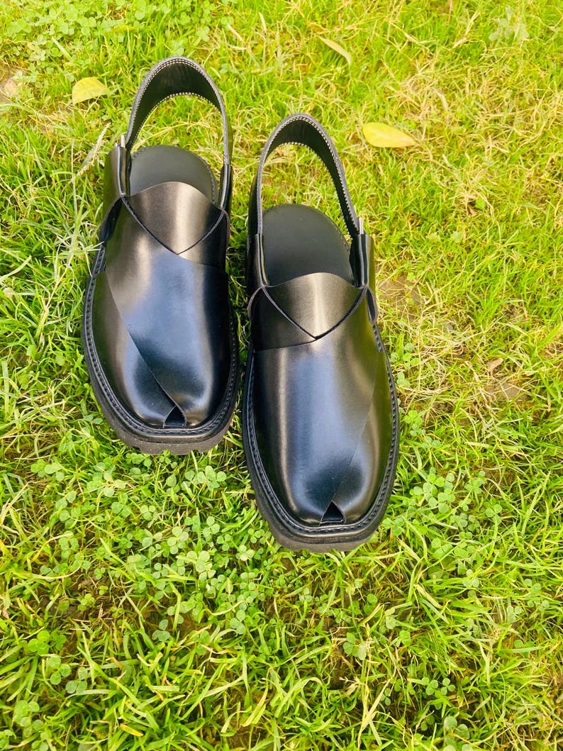 May include: A pair of black leather sandals with a strap across the top and a pointed toe. The sandals are on a green grassy surface.