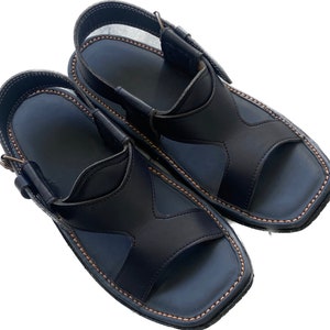 Könnte beinhalten: Ein Paar schwarze Ledersandalen mit geschlossener Zehenpartie und offenem Absatz. Die Sandalen haben eine braune Nahtdetail an den Rändern.