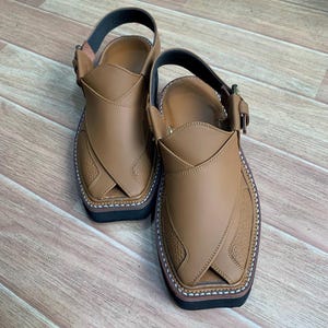 Op de afbeelding: Een paar bruinleren Peshawari-sandalen. De sandalen hebben een bovenwerk met textuur, een enkelband en een zwarte zool. De randen zijn gestikt met witte draad. De sandalen worden gepresenteerd op een houten oppervlak.